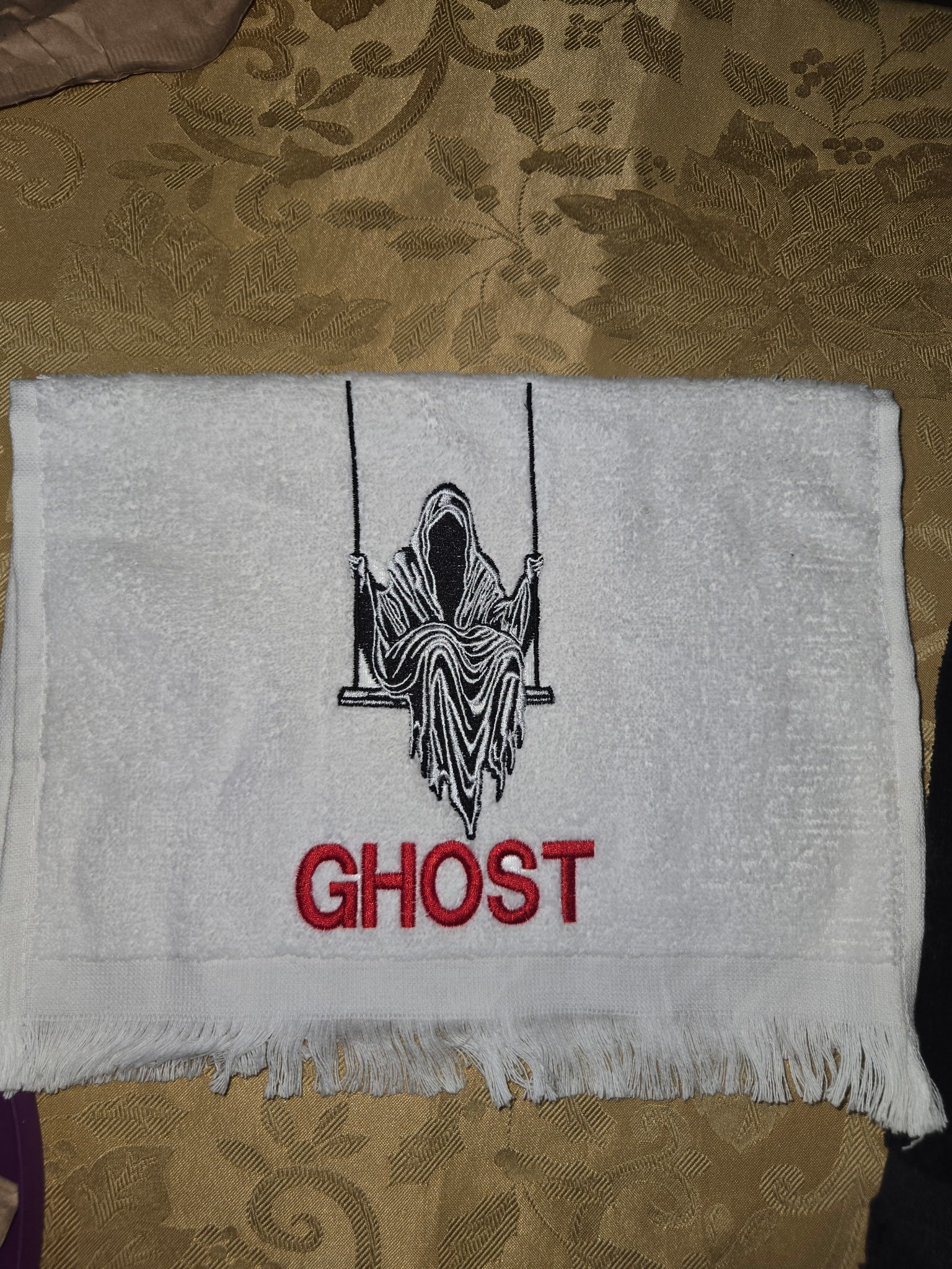 White fringed towel embroidered with a reaper on a swing and 'GHOST' lettering.