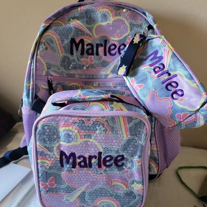 Matching pastel rainbow-and-cloud backpack, lunchbox, and pouch, each embroidered with the name 'Marlee' in purple.