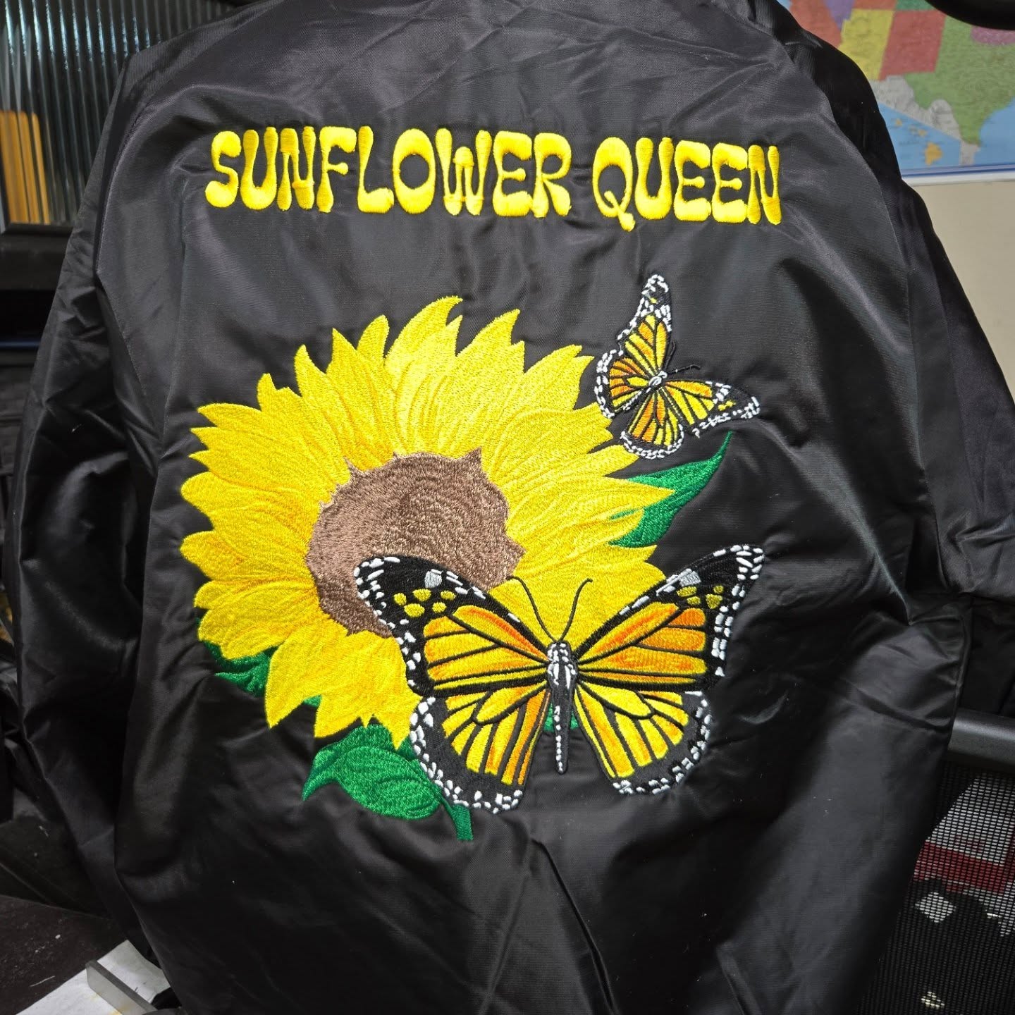 Black satin jacket with a large embroidered sunflower, monarch butterflies, and 'Sunflower Queen' lettered across the back in gold.